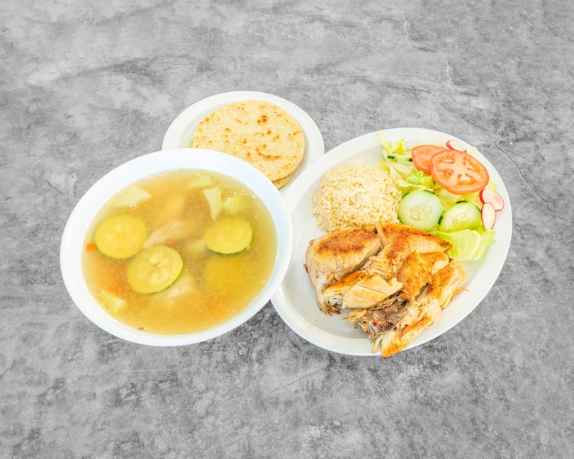 Grilled chicken with rice, salad, soup, and tortillas on a gray background.