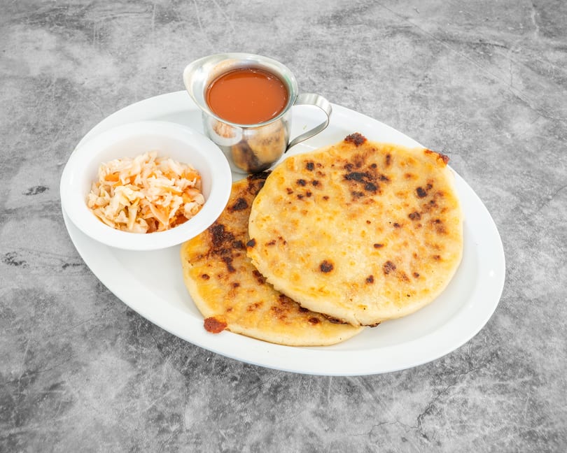 Two pupusas with curtido and red sauce on a white plate against a gray background.